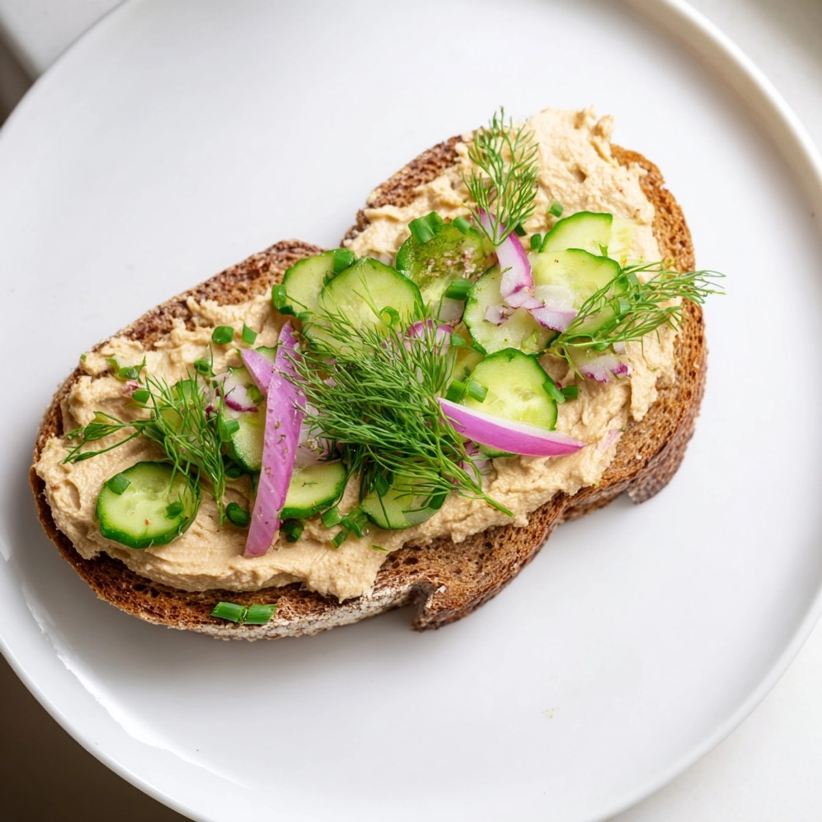 Tosty z chleba żytniego z wegańską pastą wędzoną, udekorowane szczypiorkiem i ogórkiem.