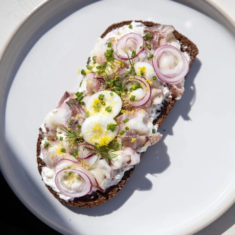 Soczysty śledź Express Cream Cheese Tartine z chrupiącą cebulą i świeżymi ziołami na bogatym ciemnym chlebie.